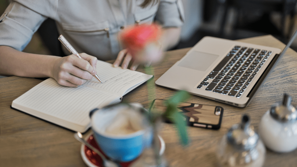 A woman writing in a notebook beside a Macbook