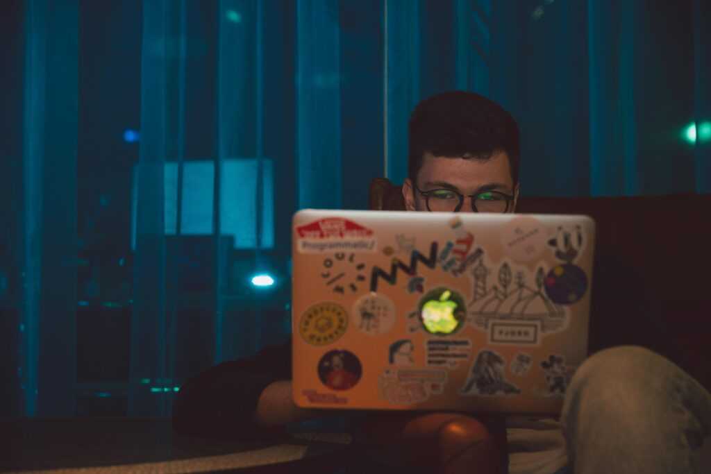 A man focused on working on his laptop with stickers at night, indoors.